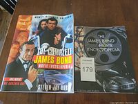 Photo showing front covers of both books side by side on wooden surface. The Complete James Bond Movie Encyclopedia book features a colorful cover with James Bond characters. The James Bond Movie Encyclopedia 2020 layout is darker with film reel and Aston Martin car.