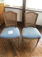 Two vintage cane back dining chairs with blue fabric cushions sitting on wood floor near window. Visible carving on legs.