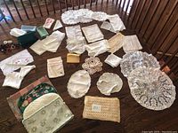 Wide view of assortment including folded cloth napkins, crocheted doilies, embroidered and quilted placemats, in white, cream, beige, green and floral patterns spread on wooden floor.