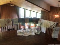 Wide shot showing various tablecloths and napkins hanging on wooden railing and folded on the floor, with multiple colors and patterns including floral, plaid, solid, and lace-trimmed.