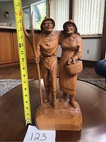 Wooden figurine of old man and woman with pitchfork and basket shown with a measuring tape to indicate height of about 16 inches, placed on a table in a room with wooden flooring and natural light from windows.