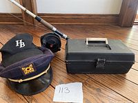 Photo of fishing rod, two hats (black baseball with HB and black captain's with skull emblem), and black tackle box.