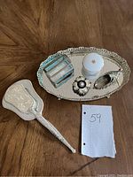 White ornate hand mirror with floral design, glass jewelry box, decorative compact, small white container with crest emblem, all on silver footed mirror tray.