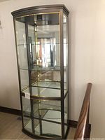 Front left side view of tall glass curio cabinet featuring brass trim, glass shelves and mirrored back.