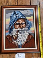 Frontal view of needlepoint depicting fisherman with blue hood and pipe, wood frame, measuring approx 15 by 20 inches