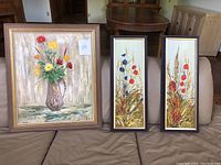 Set of 3 original paintings with wood frames shown together on sofa. Includes one larger painting and two narrow vertical paintings.