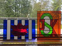 Photo showing two stained glass panels placed side by side, one with red maple leaf on blue and white background and the other with green and orange letter design.