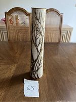 Photo of the tall thin pottery vase with dancing tree design on a wooden table with cane-back chairs in background. White paper with number '63' is placed in front of the vase.
