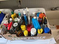All cleaning products arranged in a box on a washer, showing various spray bottles, liquid containers and cans, with some opened and some unopened items.