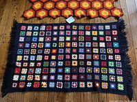 Top view of two different patterned vintage Afghan knitted blankets laid on wooden floor; one with small multicolor squares on black background with fringes, the other with large hexagonal motifs in yellow, red, and dark brown.