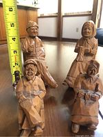 Four hand carved wooden figurines displayed on wooden floor with measuring tape showing approximate height around 3-4 inches.