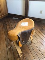 Side view of vintage leather saddle chair showing tan leather seat and darker leather sides with tassels, wooden legs, and metal stirrups.