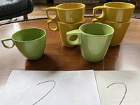 Front and side view of six yellow and two green vintage melamine tea cups placed on a wooden table, showing stacking design and geometric shaped handles.