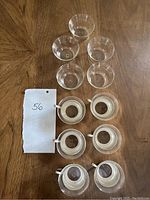 Top view of 5 clear cylindrical glasses and 6 Mainz Jena-Glas glass coffee and tea cups with metal handles on a wood surface, with handwritten note marked '56'.
