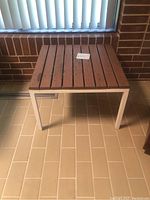 Side view of square outdoor table showing wooden slat top and metal legs on tiled floor against brick wall.