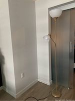 Gold metal floor lamp standing against a white corner wall with two lamp heads - a large upward-facing bowl-shaped frosted glass shade and a smaller side adjustable frosted glass shade. The lamp has a slim round base and a power cord visible on wooden flooring.