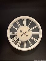 Front view of white round clock with Roman numerals and decorative hands against black wall.