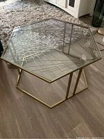Hexagonal table with glass top and gold metal geometric frame base in room setting on wood floor.