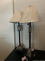 Two black table lamps on a black surface, one shade covered with plastic showing green shading.