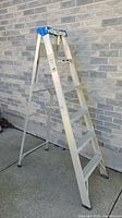 Side angle view of 6-foot aluminum step ladder showing the five steps, aluminum frame, and blue top cap against a brick wall background.