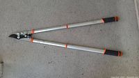 Full view of the tree pruner laid flat on concrete showing the aluminum handles, black blade, orange locking clips and grips.