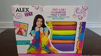 Box front showing brand ALEX DIY, product name Knot A Quilt, dimensions 107 cm x 137 cm (42 in x 54 in), and image of completed quilt held by a girl.