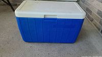 Closed view of large blue and white Coleman cooler showing textured blue exterior and white lid with latch.