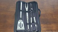 Full view of the BBQ utensil set in black carrying case showing spatula, fork, and tongs secured with elastic straps.