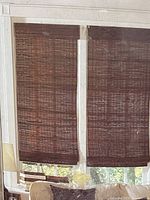 Two brown bamboo woven window shades hanging in a window, partially rolled down, showing the woven texture and light filtering properties.