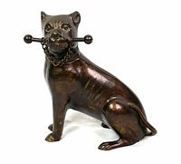 Bronze dog figurine sitting, holding a rod with round ends in its mouth, chain around neck, side view
