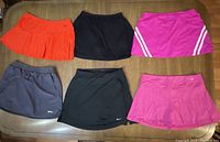 Six women's tennis skirts arranged flat on a surface showing various colors and styles including orange, pink, black, and grey.