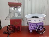 Popcorn machine and cotton candy maker shown side by side on a red table surface against a white background.