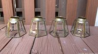Four vintage hexagonal glass light covers with brass frames arranged on wooden surface, showing tapered shape and round top openings.