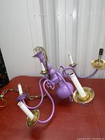 Purple painted brass chandelier with five candle arms, gold cup accents under sockets, and hanging chain shown from two angles on a red surface.