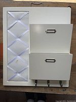 Full view of the white wall-mounted mail and keys sorter message board showing two metal compartments with label holders, three black hooks, and ribbon section for holding mail or notes.