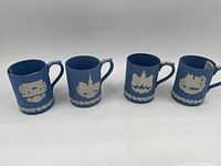 Front view of four blue Jasperware Wedgwood mugs showing different historic white bas-relief designs and leaf border near bottom.