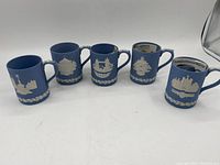 Photo of 5 Wedgwood blue Jasperware mugs in a row showing the varied white relief cityscape and architectural decorations on the front sides and planar blue background.