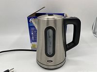 Front view of Oster stainless steel electric kettle showing its shape, handle, spout and water level indicator, plugged in and on a white surface with its original box in background.