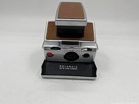 Front view of Polaroid SX-70 Land Camera showing red shutter button, viewfinder aperture, and model label on black base plate.