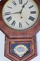 Close-up photo of the clock dial showing Roman numerals, hour and minute hands, and part of the wooden octagon case and lower door with decorative glass.