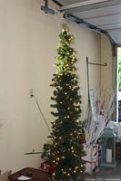Artificial pencil Christmas tree standing assembled in garage against wall, lit with white string lights, topped with a decorative star, some visible bell ornaments hanging along the branches