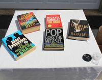 Five assorted thriller books arranged on a white tablecloth outdoors, visible titles and covers, showing a mix of hardcover and softcover.