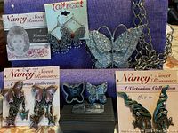 Photo showing multiple butterfly-themed jewelry pieces arranged on a purple fabric background, including a large silver-tone butterfly pendant necklace, pairs of purple/gold Victorian clip-on earrings, silver-tone lace pierced earrings, blue/black butterfly studs in a box, turquoise clip-on earrings, and a miniature jewelry box.