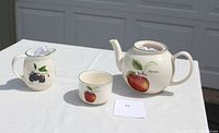 Photo showing milk pitcher with plum design, sugar bowl with apple design, and small cup with peach design on white cloth background.