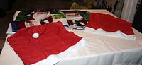Two red fabric Santa hat chair covers with white pom-poms on top and white trim, laid flat on a table.