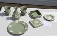 Overall view of green Wedgwood jasperware set on a white table, showing two urns, one bottle-shaped vase, two plates, and one square box with lid.