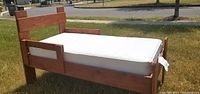 Side view of hand-crafted wooden toddler bed with white mattress outdoors on grass.
