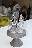 Front and side view of the pewter cruet stand with four glass cruets labeled for condiments. One cruet lacks a stopper.