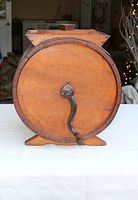 Front view of vintage wooden butter churn showing circular wooden barrel with metal hand crank and wooden feet on a white table.