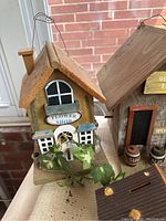 Photo showing two wooden decorative bird houses including a flower shop and a general store style, placed indoors with brick wall background.